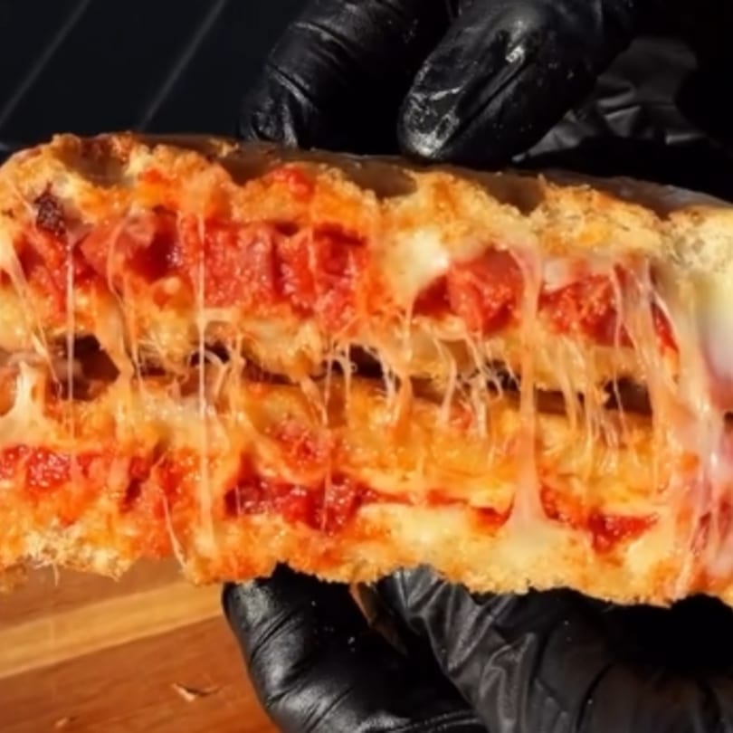 Gloved hands pull apart a cheesy, pepperoni-filled pizza sandwich, stretching the melted cheese.