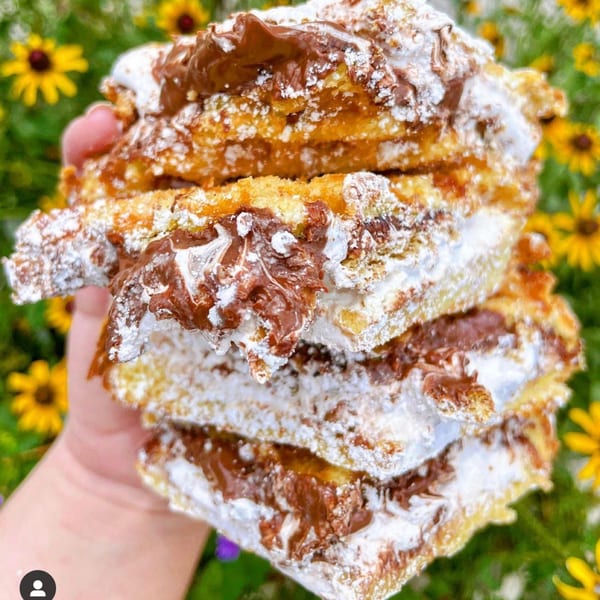 A hand holds a stack of golden-brown churros, drizzled with chocolate and powdered sugar, against a floral background.