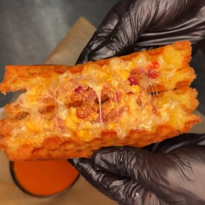 Hands in black gloves hold a cheesy waffle sandwich with bacon and corn, dripping cheese over a red sauce.