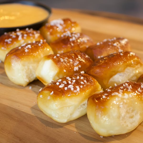 Golden brown pretzel bites with salt crystals on a wooden board, with a bowl of cheese dip.