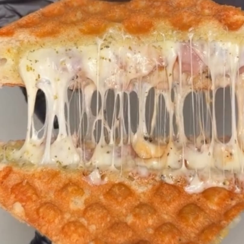 Close-up of a golden-brown waffle sandwich with melted cheese and ham stretching between the halves.