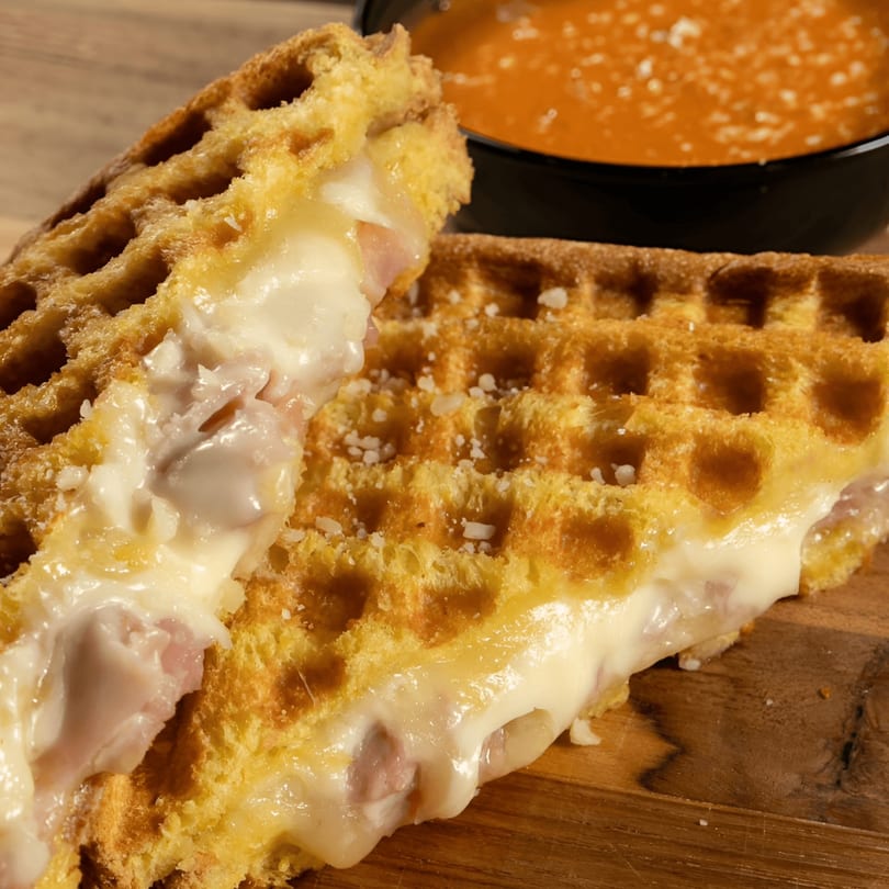 Golden waffle sandwich filled with melted cheese and ham, served with a bowl of tomato soup.