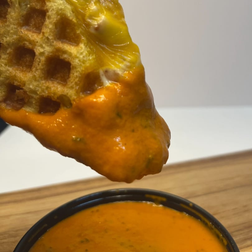 A golden waffle fry, covered in melted cheese, being dipped into a bowl of orange sauce.