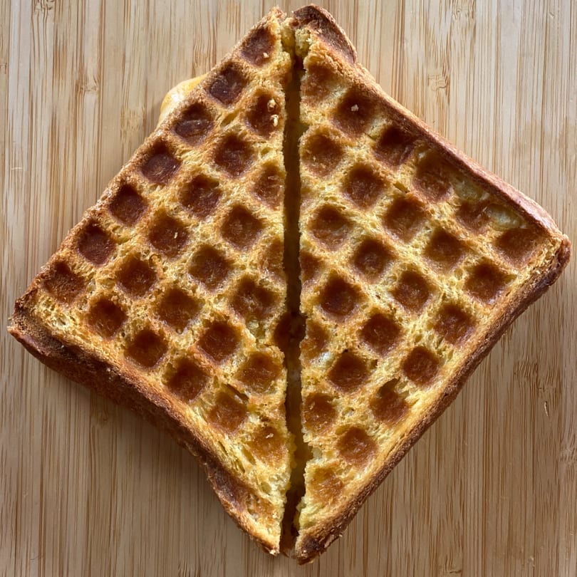 A golden-brown waffle-pressed sandwich, cut in half diagonally, rests on a light wooden surface.