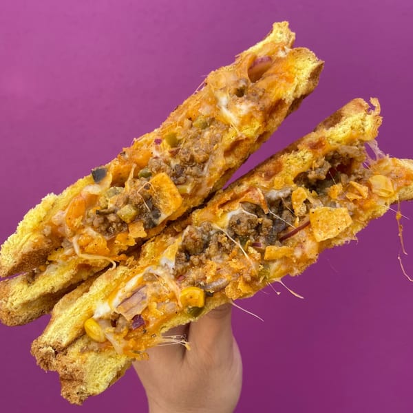 Hand holding a golden-brown, cheesy sandwich filled with ground meat, corn, and red onion.