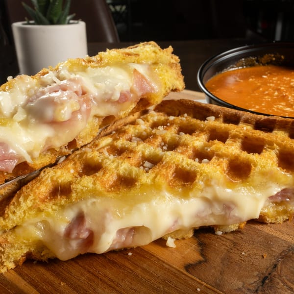 Golden waffle sandwich filled with melted cheese and ham, served with a side of tomato soup.