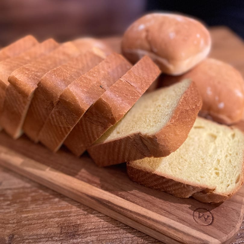 Sliced white bread and dinner rolls on a wooden cutting board.