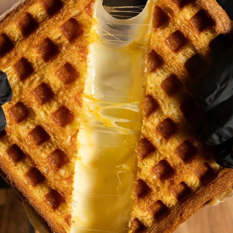 Golden-brown waffle sandwich pulled apart, revealing gooey, melted cheese stretching between the halves.