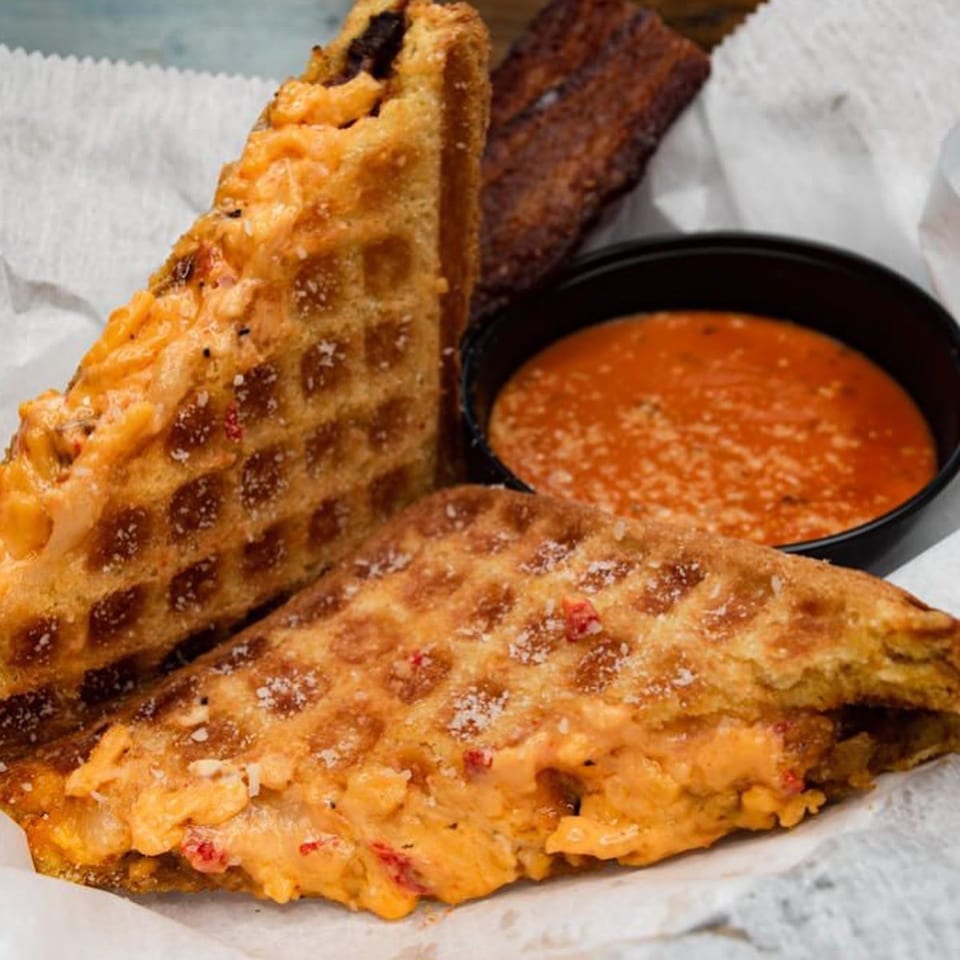 Two waffle-pressed grilled cheese sandwiches with a side of tomato soup and a piece of bacon.