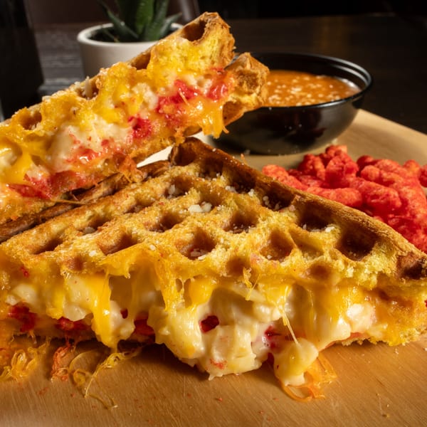 Cheesy waffle sandwich with red flakes, served with a side of tomato soup and crunchy red snacks.