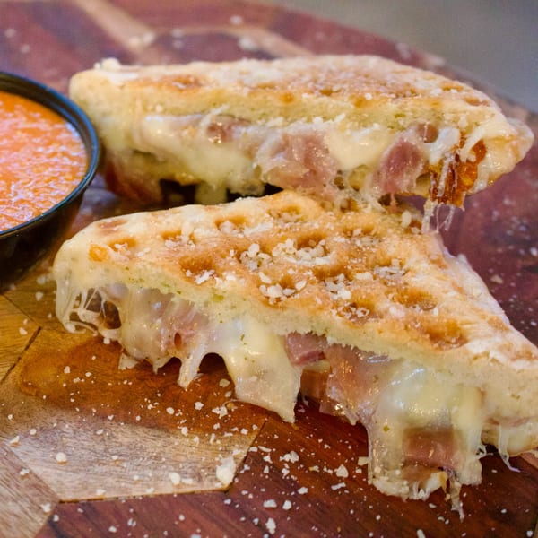 Two cheesy waffle sandwiches with ham and melted cheese, served with a side of tomato soup.