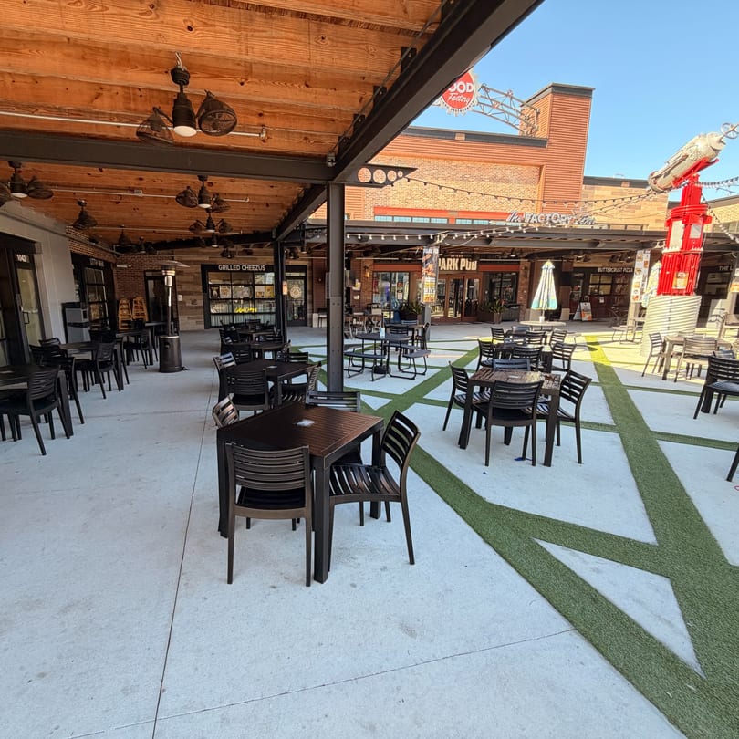 Outdoor dining area with black tables and chairs, a wooden ceiling, and various restaurant storefronts.