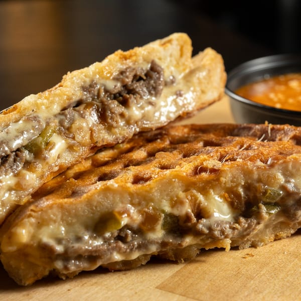 A grilled sandwich, cut in half, revealing melted cheese, meat, and vegetables, with a side of dipping sauce.